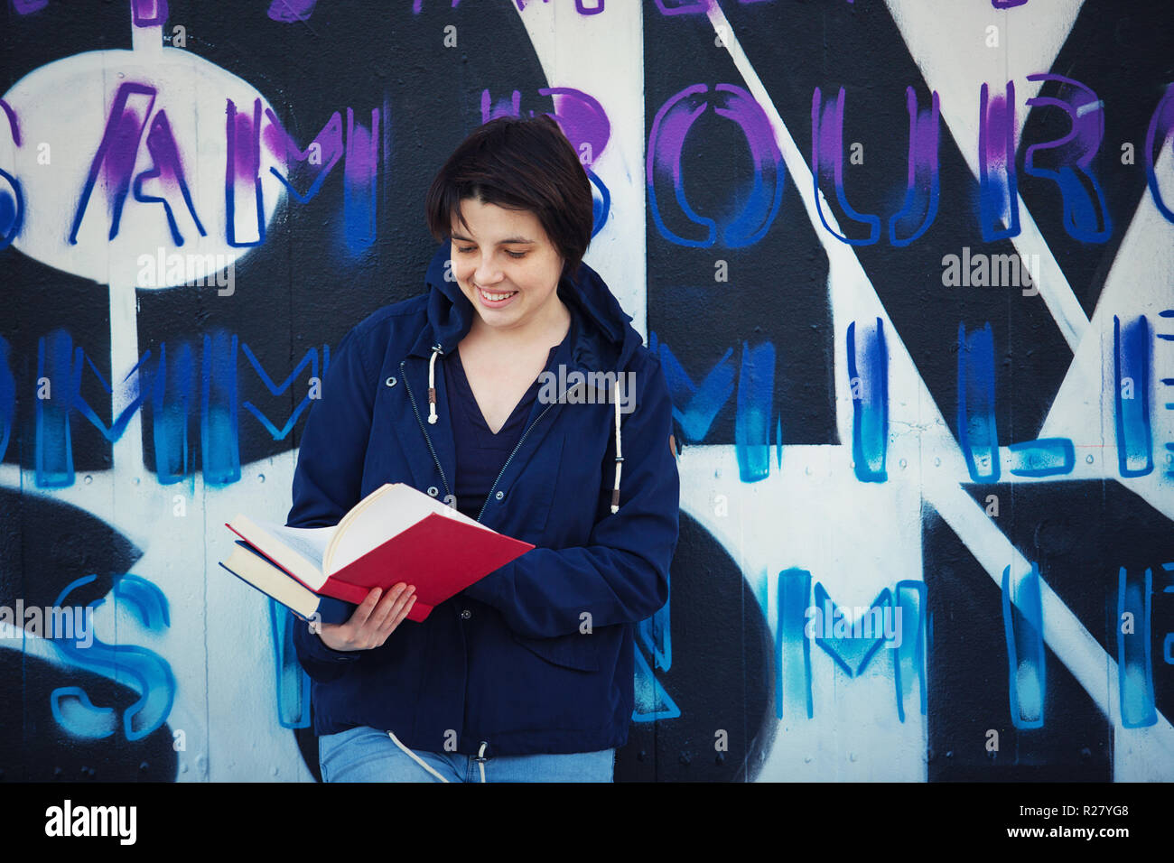 Casual mujer feliz libro de lectura del estudiante, el estilo de vida al aire libre retrato reclinado hacia atrás sobre Graffiti Wall fondo. Concepto de educación, Street art. Foto de stock