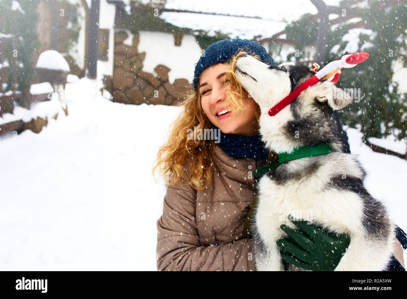 Atractiva mujer caucásica abrazos gracioso malamute perro vistiendo santa queridos astas de navidad. Rizado mujer sonriente divirtiéndose con cachorro huskie en año nuevo. El Pet es el mejor regalo. Doggy lamer cara de la niña. Foto de stock