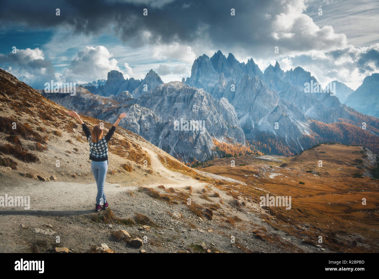 Mujer joven con los brazos levantados y las majestuosas montañas al atardecer en otoño en Dolomitas, Italia. Paisaje con la niña alegre, espectacular cielo con nubes, oh Foto de stock