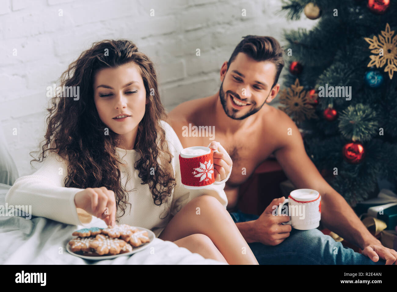 Rizado joven mujer con taza de café tomando ginger cookie mientras su novio sentado cerca de árbol de navidad en casa Foto de stock