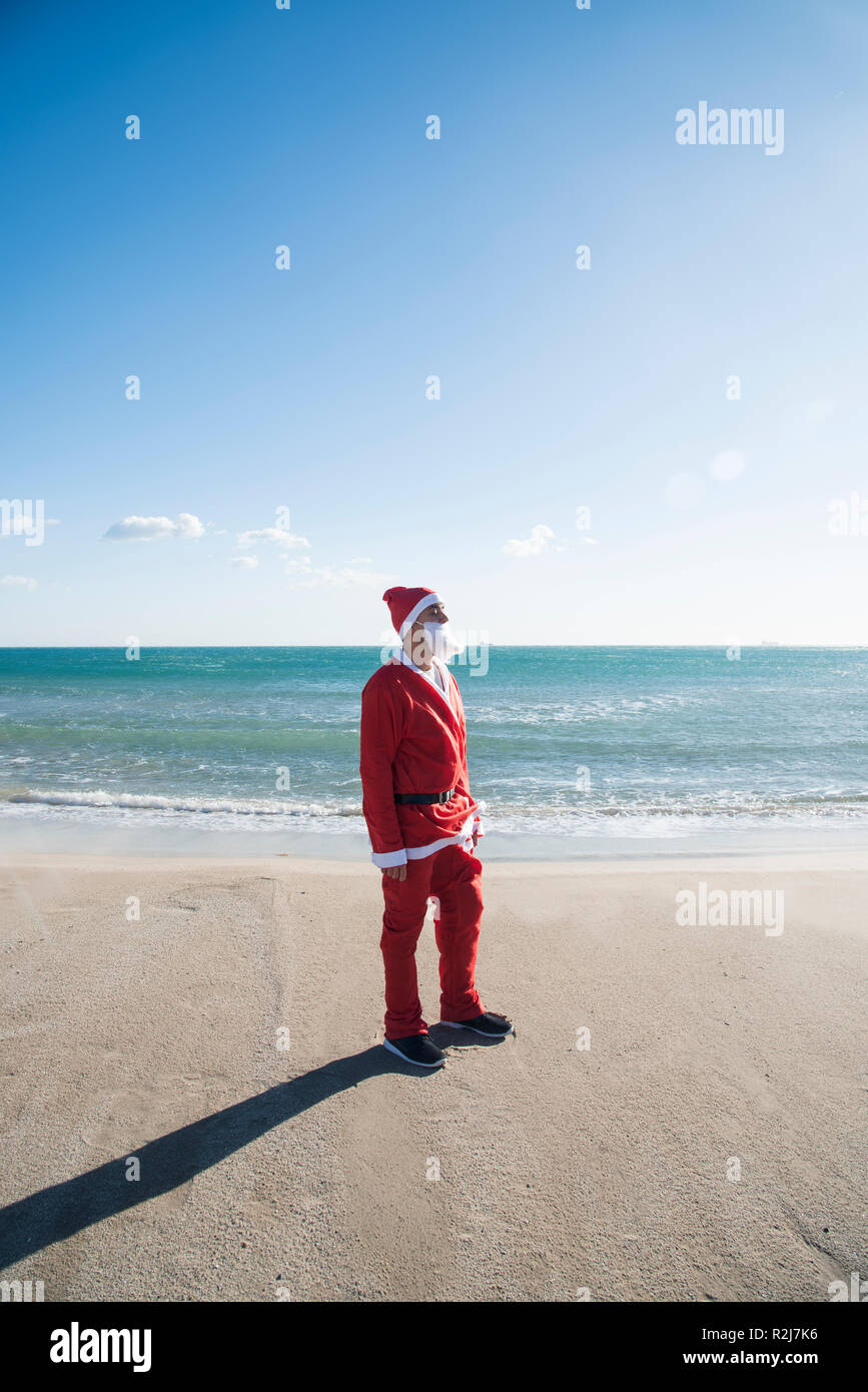 Joven vistiendo un traje de Papá Noel de pie en la playa de frente al sol, con el mar de fondo Foto de stock