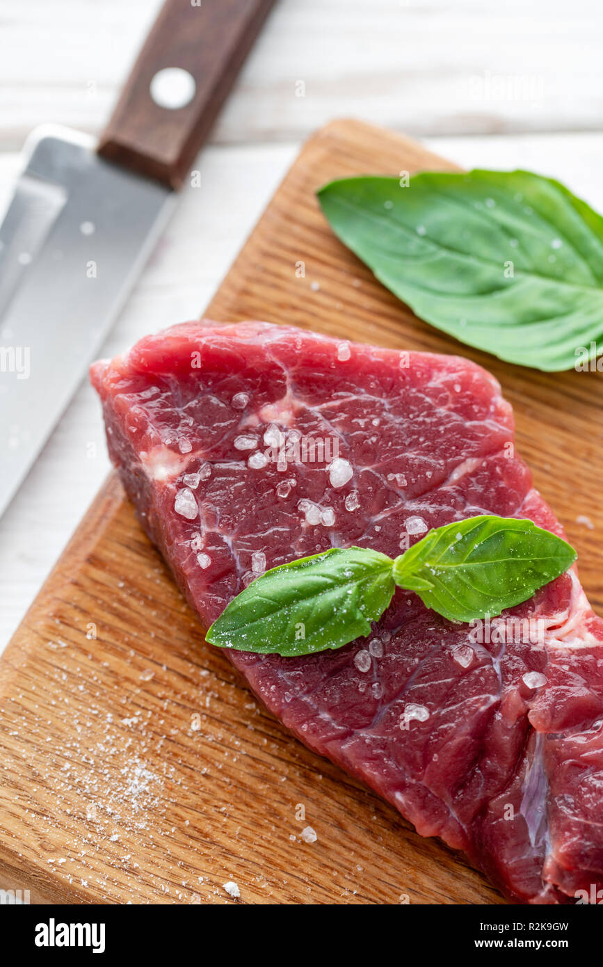 La carne adobada con sal, pimienta y condimento Albahaca hoja. Por la mañana en la cocina. Filete de carne de vacuno fresca cruda sobre una tabla para cortar madera. Fondo de madera blanca, vista superior, copia el espacio, luz natural Foto de stock