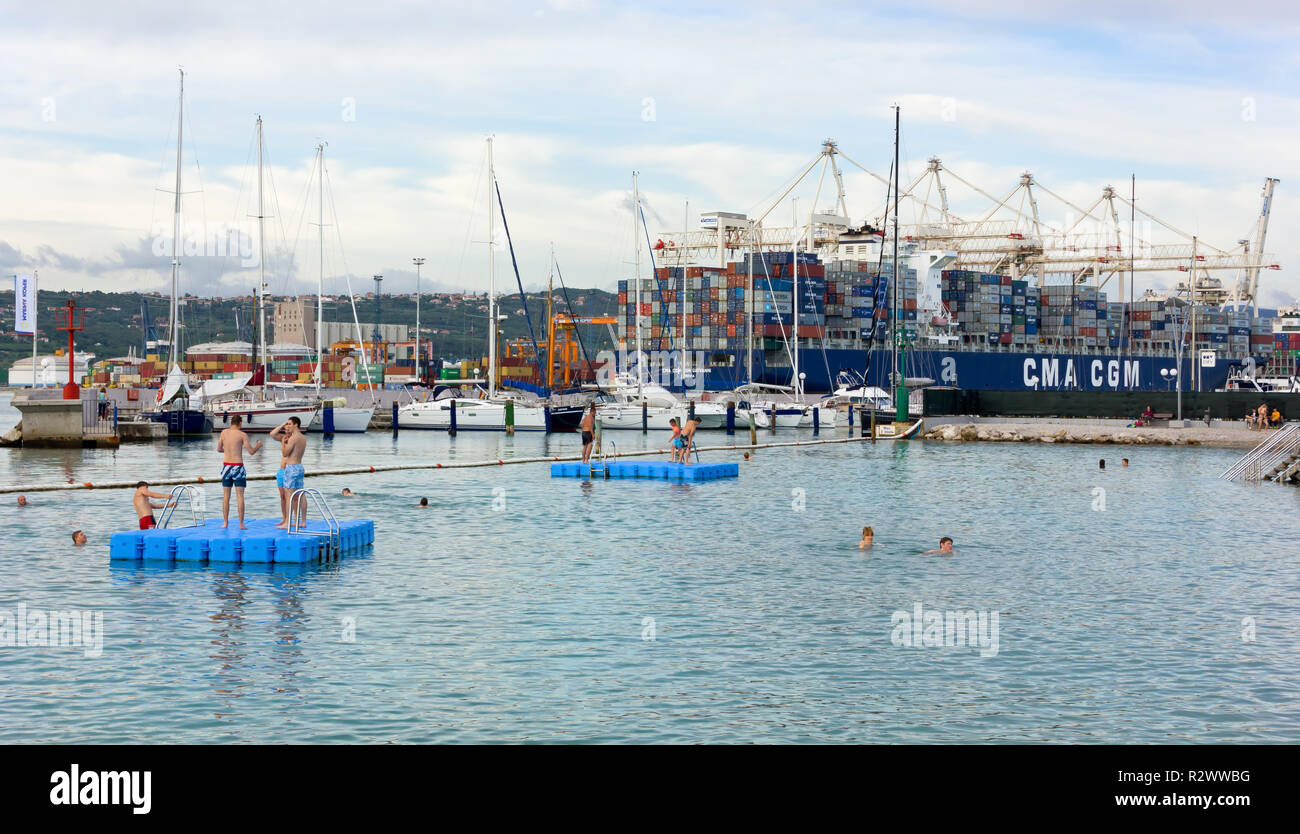KOPER, Eslovenia - Julio 24, 2014: unos pocos bañistas en las aguas antes del puerto, con un gran buque portacontenedores en el fondo Foto de stock