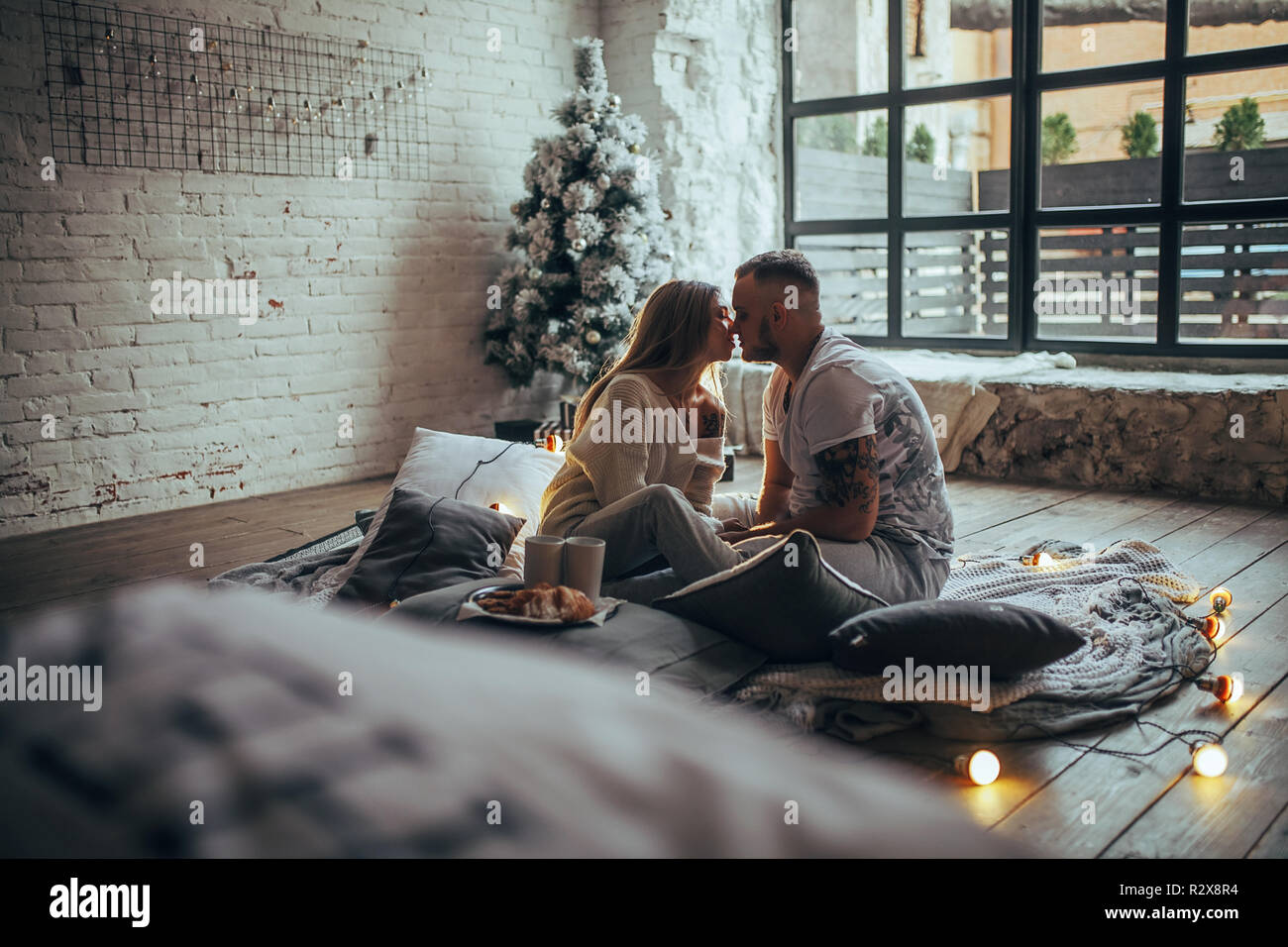 Pareja joven en el amor está sentado en el piso y besar junto a dos tazas y placa con cookies sobre fondo de árbol de Navidad bombillas incandescentes, bric Foto de stock