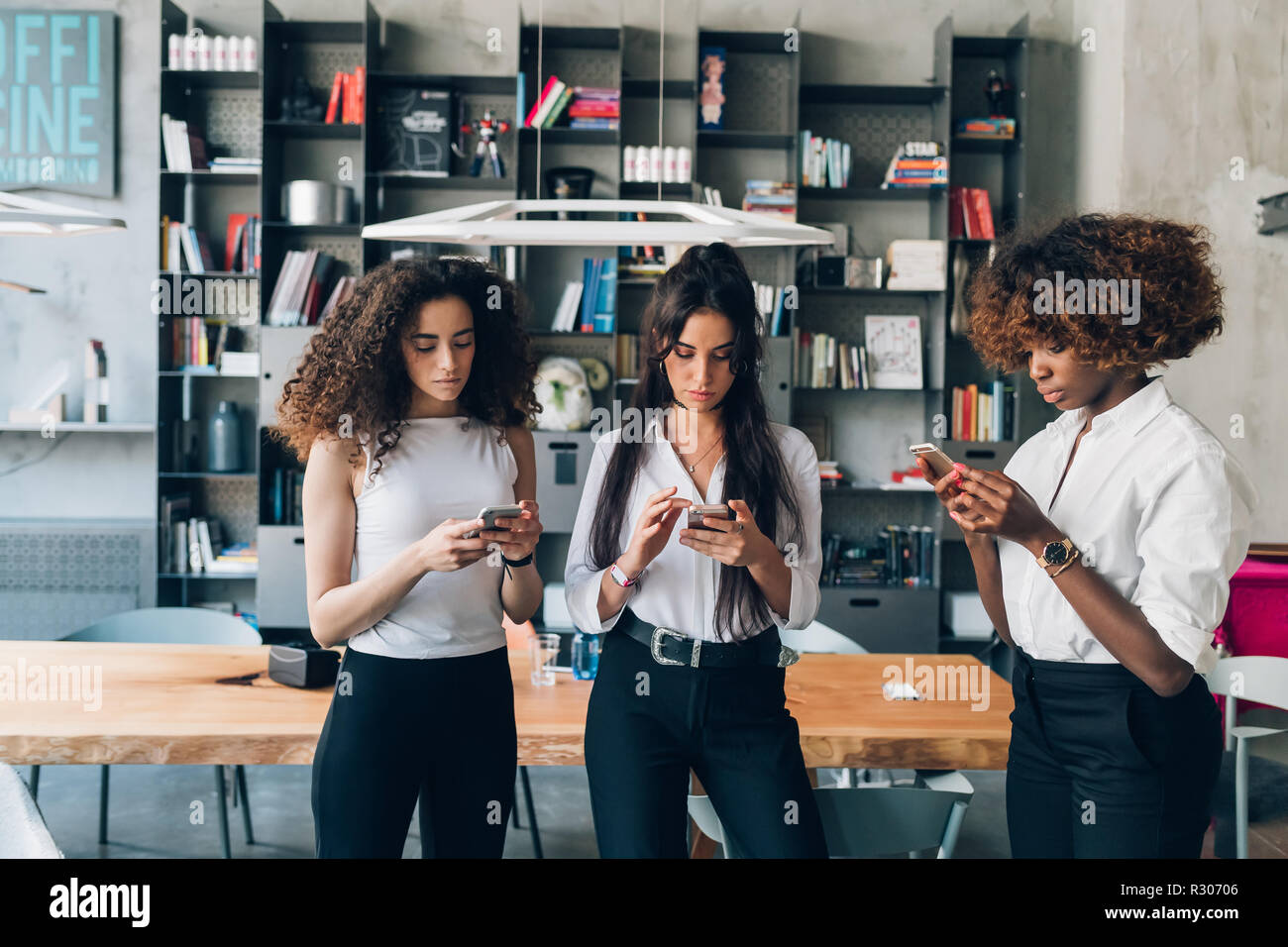Tres jóvenes mujeres equipo empresarial interiores utilizando smart phone - el trabajo a distancia, phubbing, concepto de negocio Foto de stock