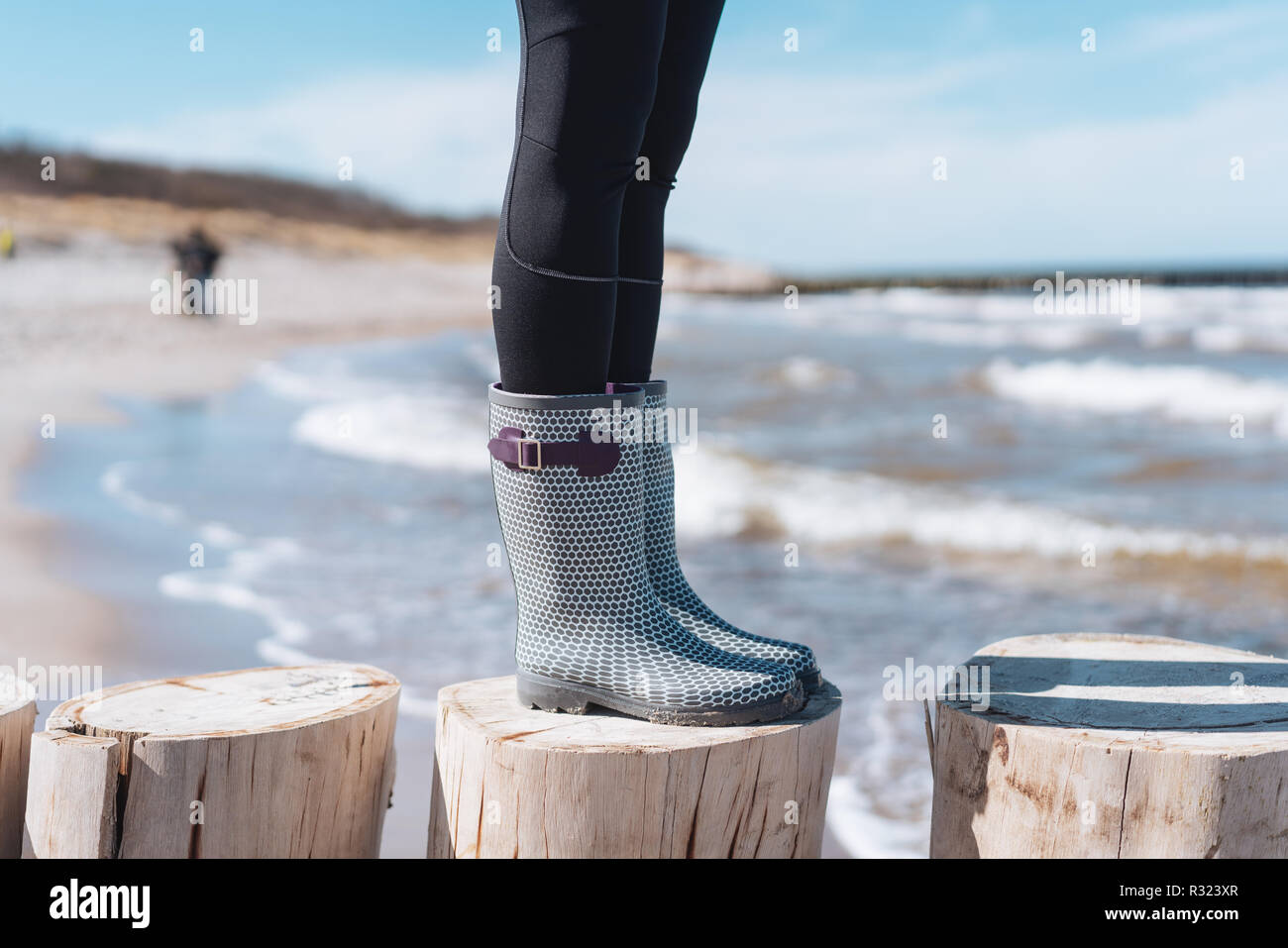 Mujer en botas de goma a la orilla del mar de pie sobre postes de madera  formando una defensa costera en una vista de cerca de sus pies con ondas  detrás Fotografía