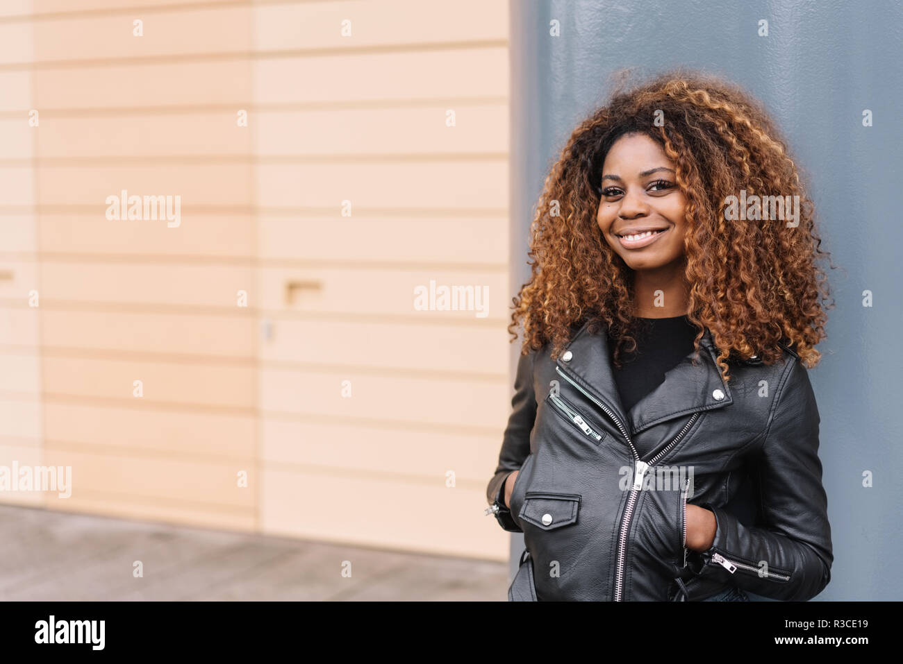 Relajado moda mujer africana en una chaqueta de cuero negro de pie apoyado contra una pared urbana con las manos los bolsillos sonriendo ante la cámara, con copia s Fotografía de