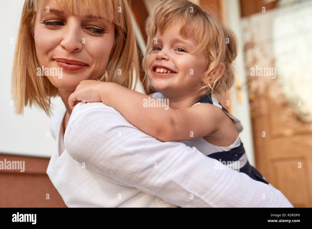 Madre sonriente llevando a su hija piggyback delante de su casa Foto de stock
