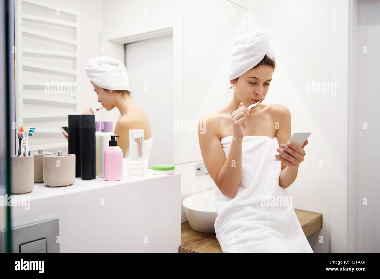 Mujer joven en el baño cepillar sus dientes mientras la lectura de mensajes de teléfono móvil Foto de stock