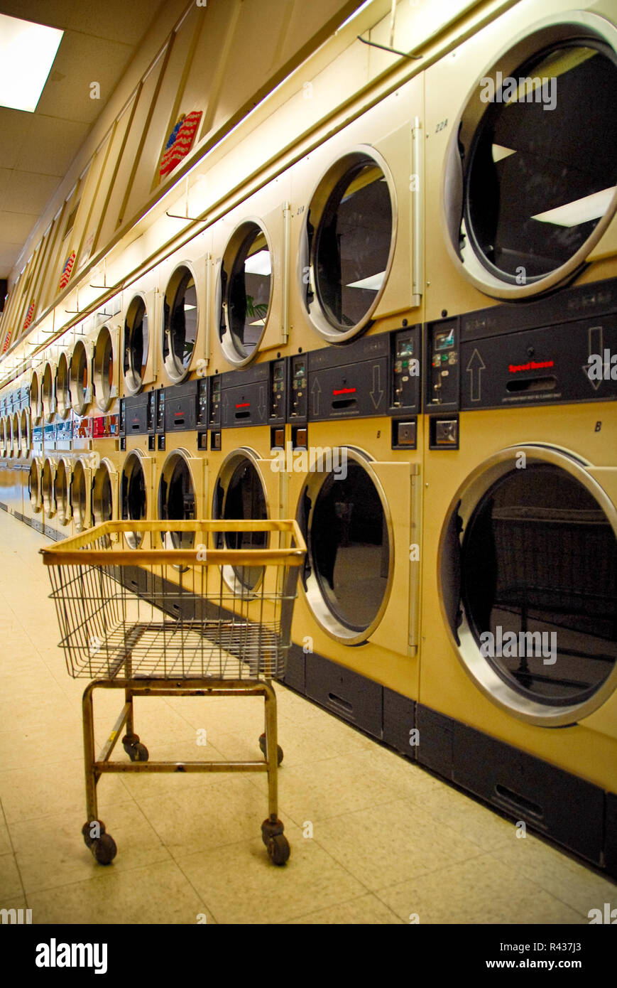 Un carro de ropa se sienta en frente de una larga línea de máquinas de lavar en una lavandería local. Foto de stock