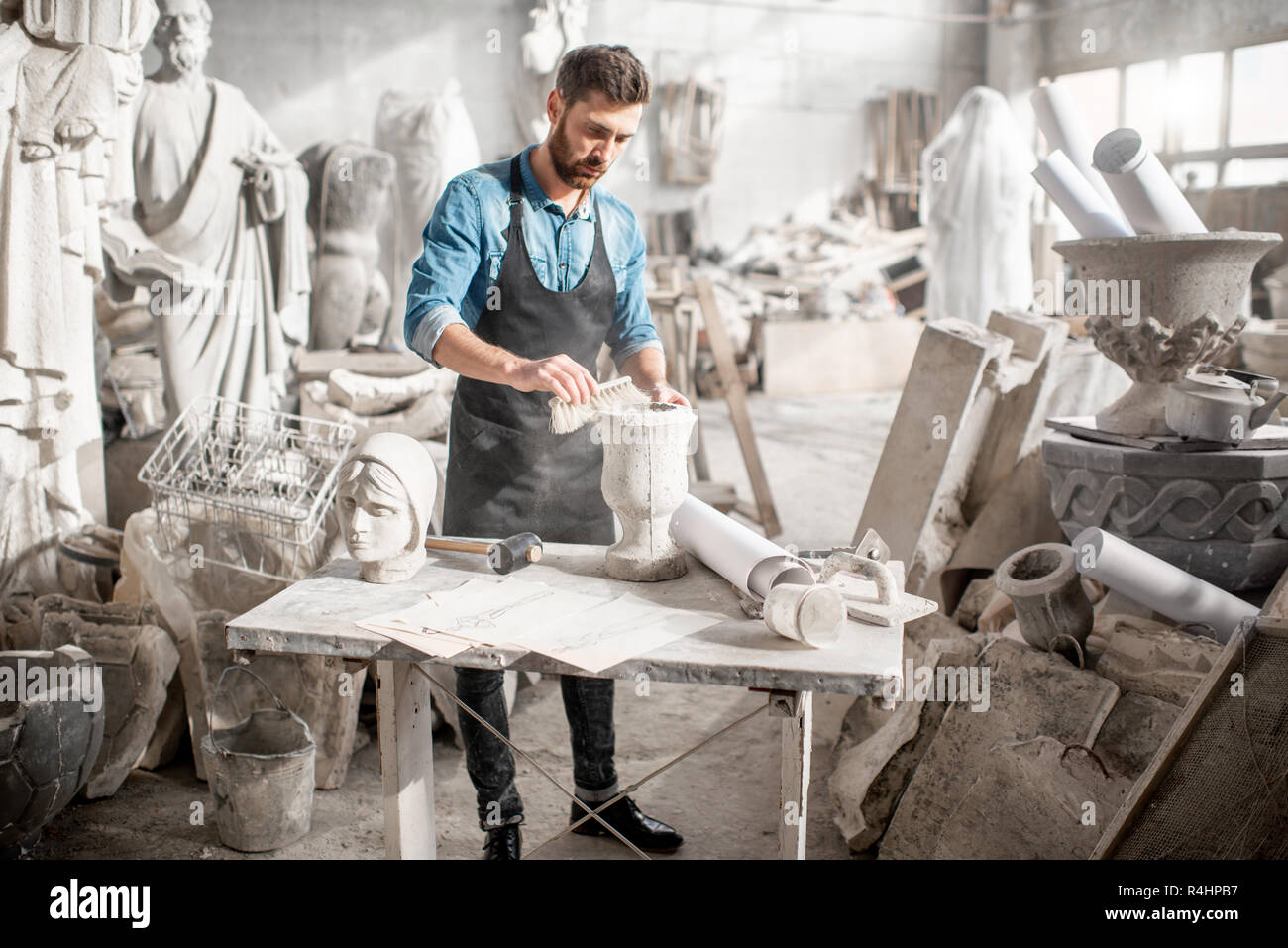 Hombre en camiseta azul y delantal trabajando en el antiguo estudio atmosférico cepillado jarrón de piedra en la mesa Foto de stock