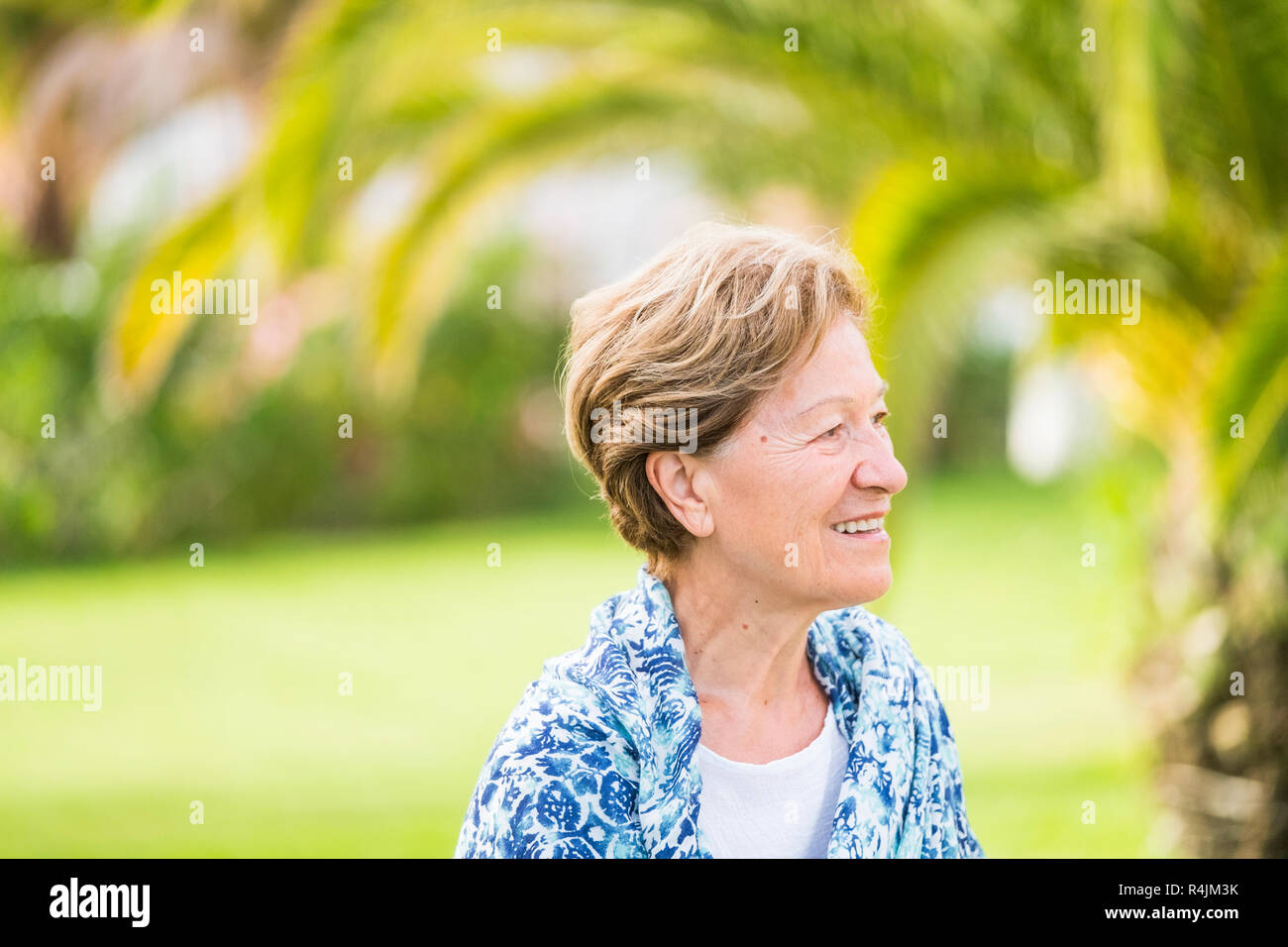 Retrato de 70 años de edad adult senior mujer sonriendo y mirando el exterior con un efecto de desenfoque bokeh de fondo verde - gente jubilada con agradable estilo c Foto de stock