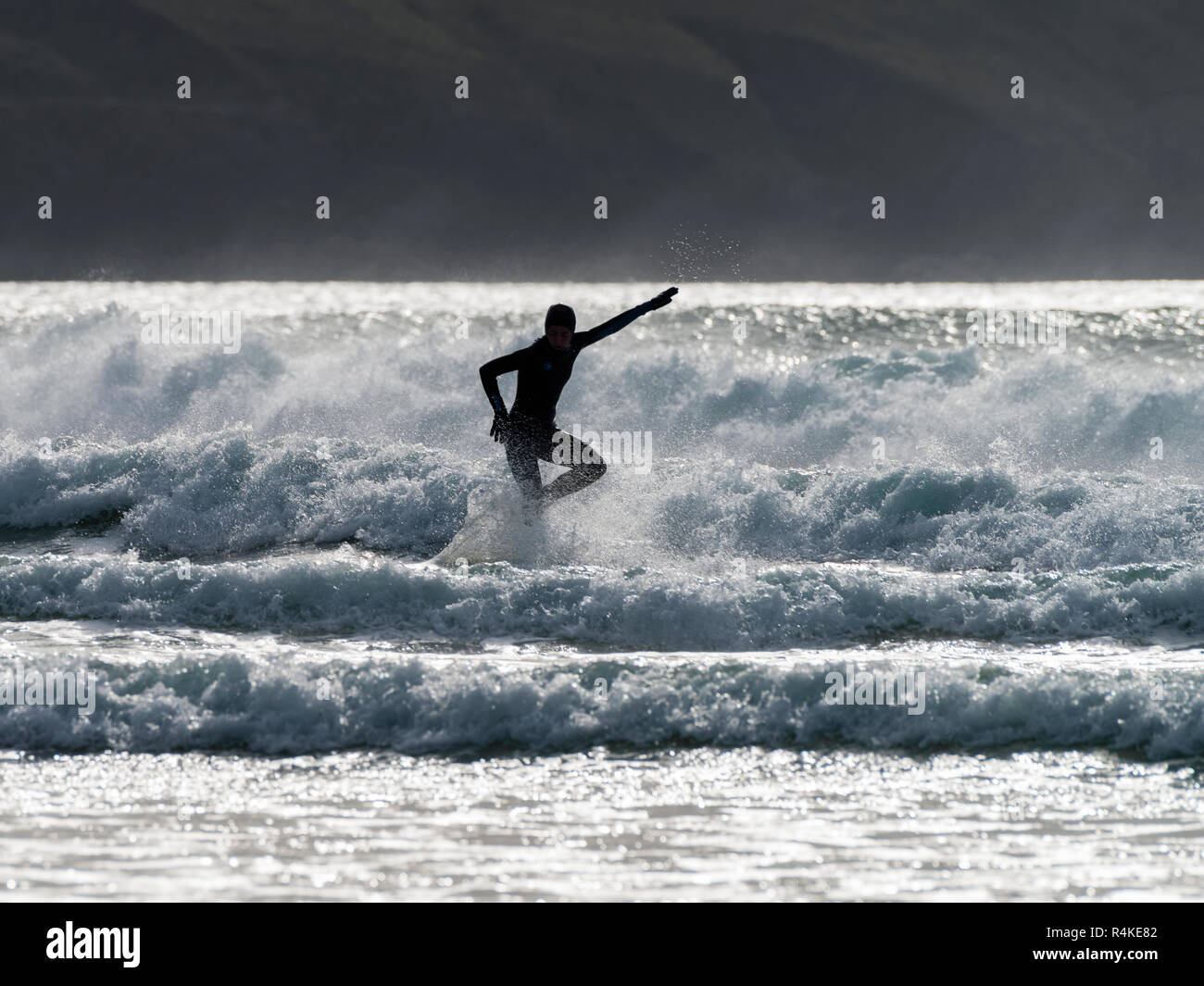 Chica surfista en neopreno golpea una pose manteniendo el equilibrio en las olas contra un telón de fondo dramáticamente iluminado. Foto de stock