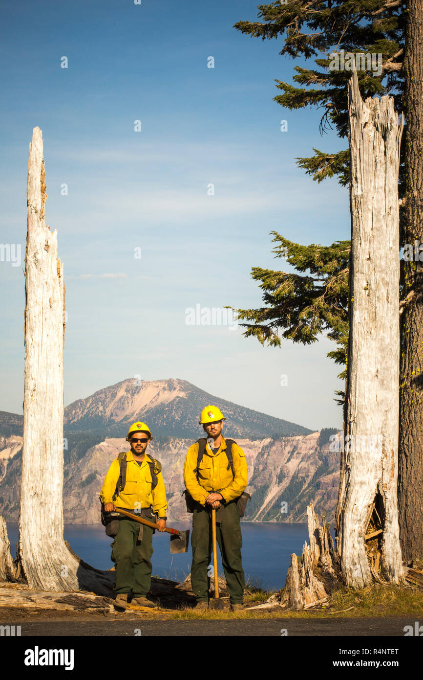Dos bomberos forestales en camisas amarillas stand con su equipo entre dos árboles muertos con un lago de montaña en el fondo. De Crater Lake, Oregón, EE.UU. Foto de stock