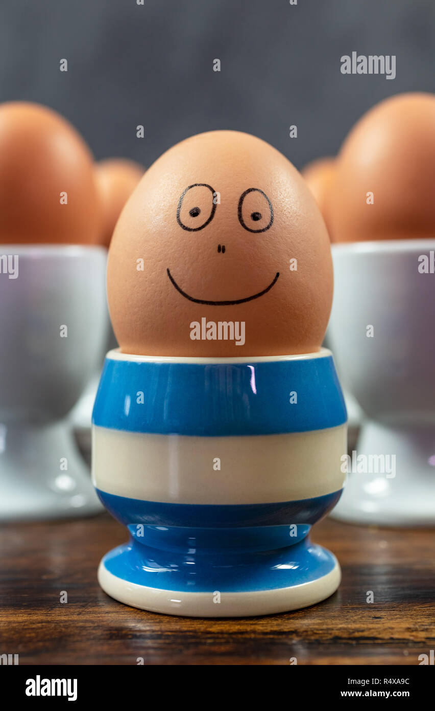 Feliz emoción concepto uno cada huevo cocido sonriente en diferentes huevera de rayas azul rodeada de huevos en tazas de huevo blanco sobre una mesa de madera Foto de stock