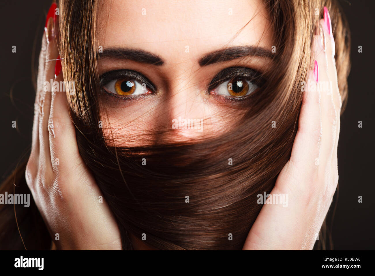 Cerrar mujer cubre el rostro por largos pelos marrones ojos marrones con maquillaje oscuro Foto de stock