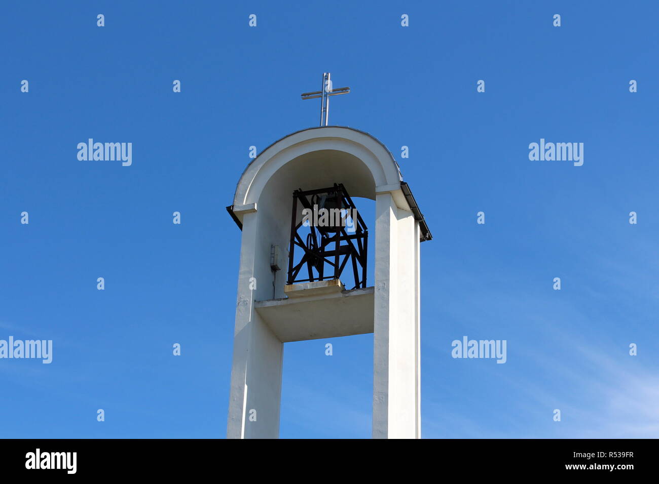 Moderna torre campanario de la iglesia de blanco con una gran campana de  metal abierto, mecanismo de techo redondeado y cruz de acero brillante en  la parte superior con el azul claro