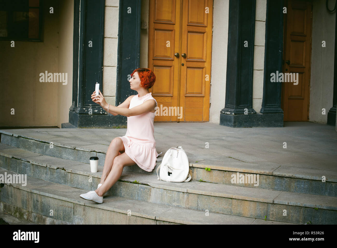 Chica haciendo selfie. mujer teñida de cabello rojo en un vestido rosado  pálido con una mochila color blanco de beber café, fotos de sí mismos en  sus teléfonos móviles con cámara, sentada