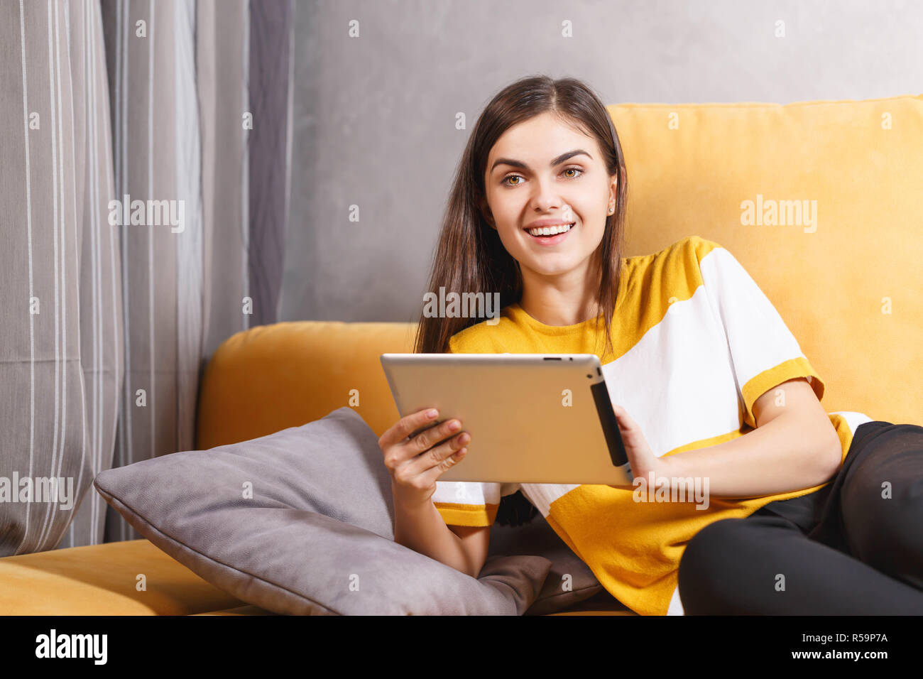Feliz niña morena de pelo largo acostado en el sofá amarillo en el salón de luz y el uso de la tecnología moderna, tableta digital, el concepto de comunicación Foto de stock