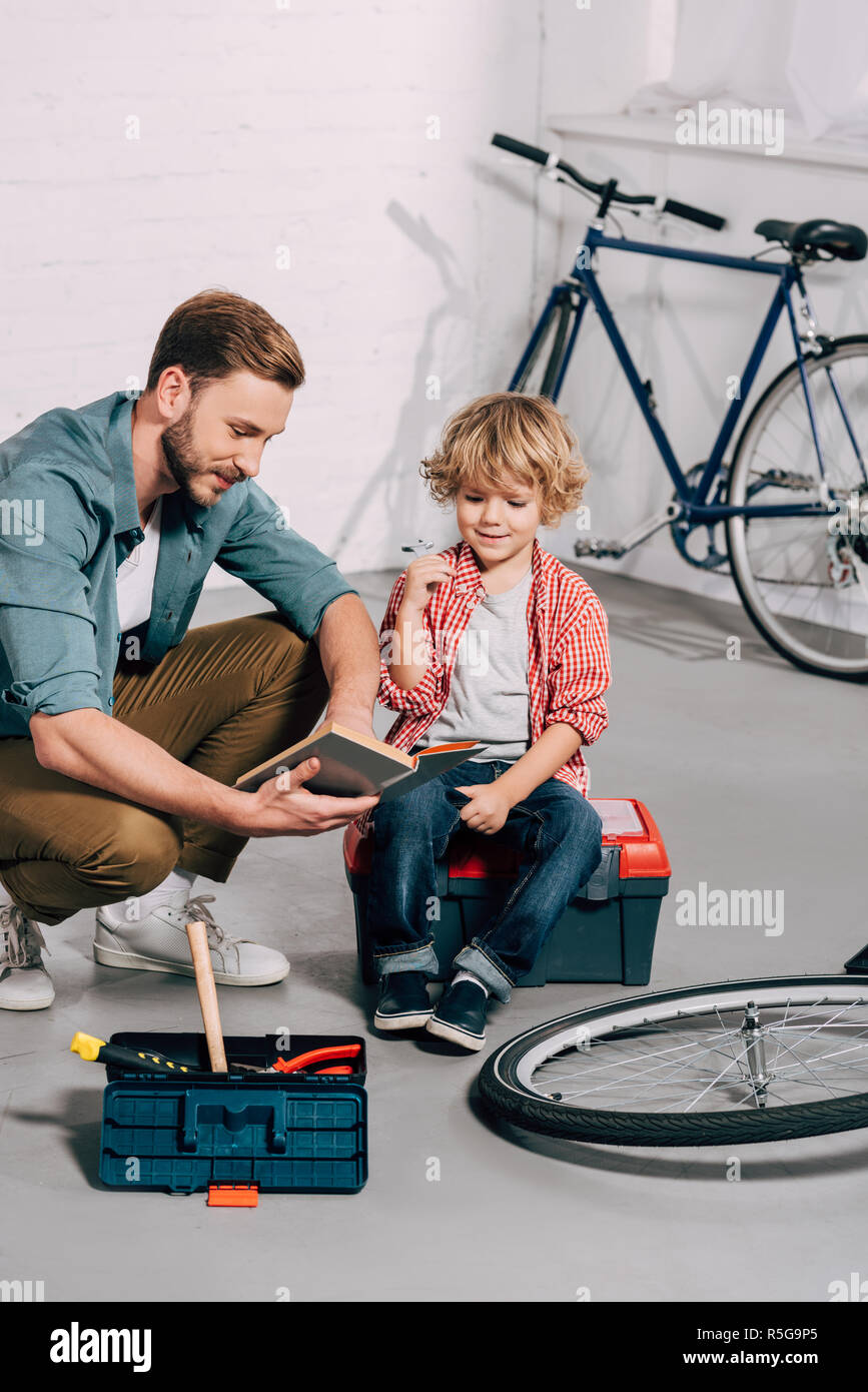 Feliz el hombre apuntando con la mano al libro a pequeño hijo sentado en la  caja de herramientas en el taller de bicicletas Fotografía de stock - Alamy