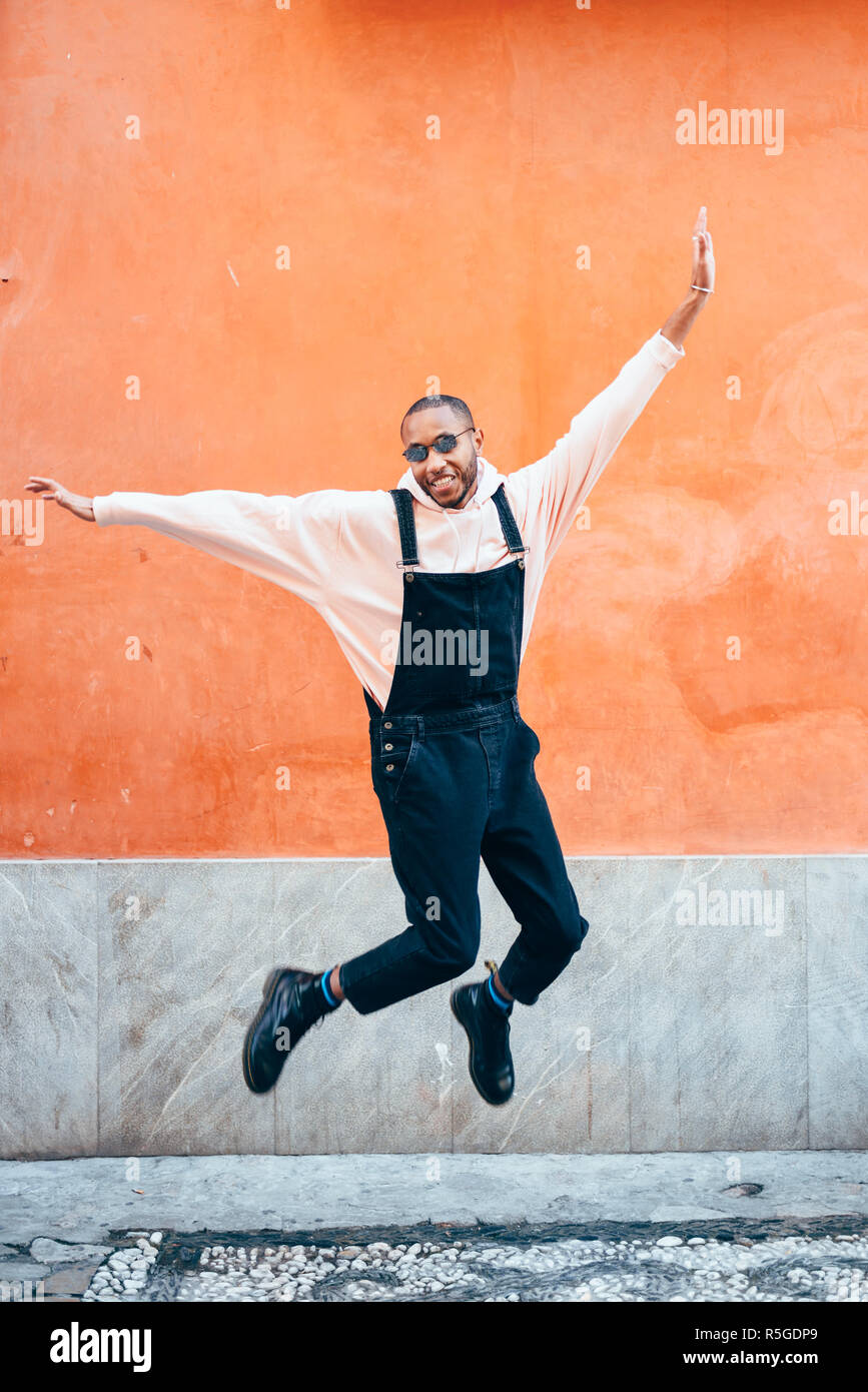 Joven negro vistiendo ropa casual saltar en el contexto urbano. Concepto de estilo de vida. África milenaria chico con pantalones de bib en el exterior Foto de stock