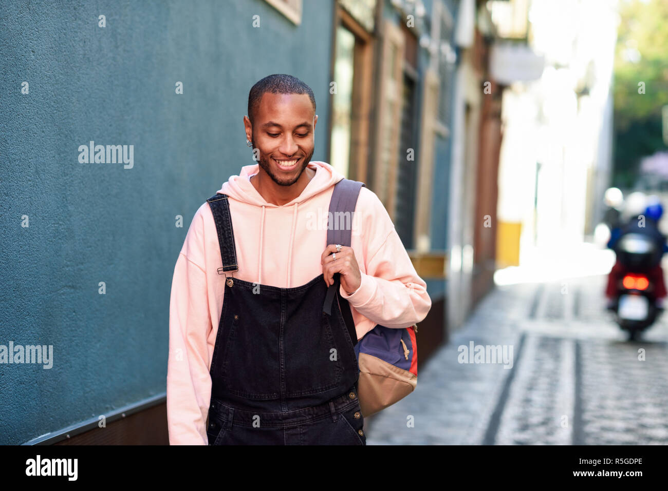 Joven negro vistiendo ropa casual sonriendo caminando por la calle. África milenaria chico con pantalones de bib en el exterior Foto de stock