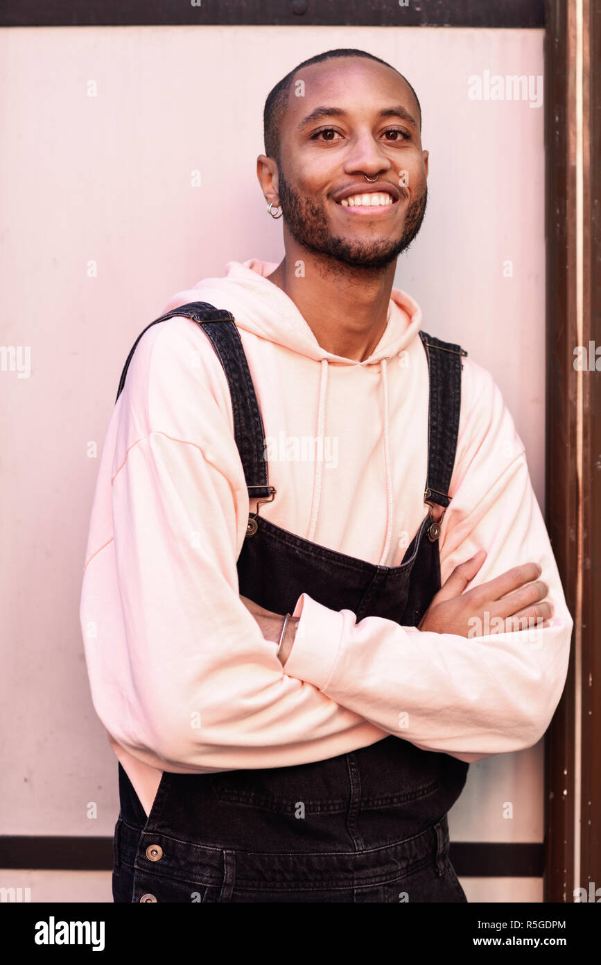 Joven negro vistiendo ropa casual sonriendo contra rosa de fondo urbano. Atractivo chico africano con pantalones de bib en el exterior Foto de stock