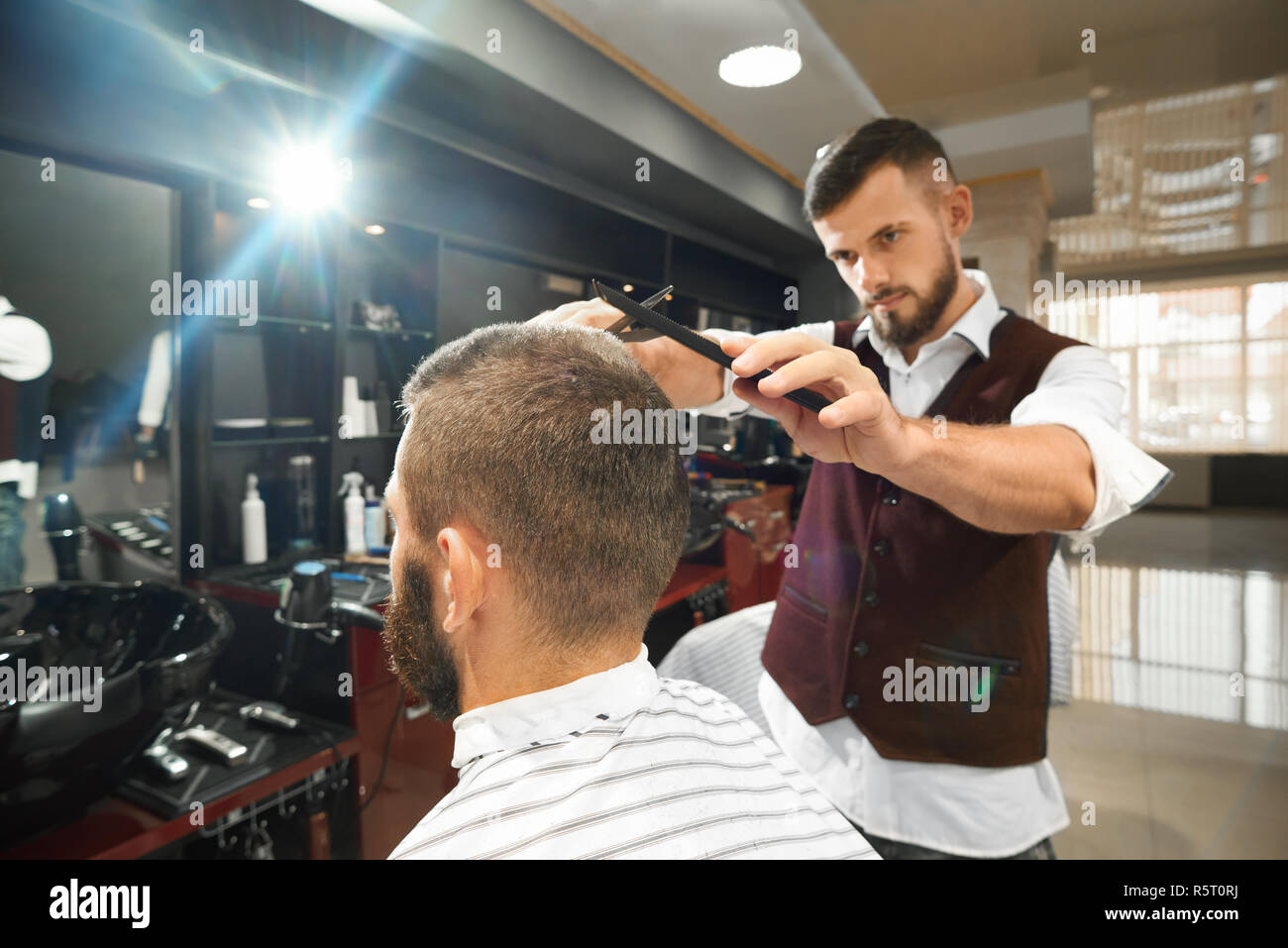 Seria brutal barber en uniforme de pie cerca de clientes hombres sentados  en una silla y haciendo elegante corte de pelo en la peluquería. Hombre  calificado con peine y recta de afeitar.