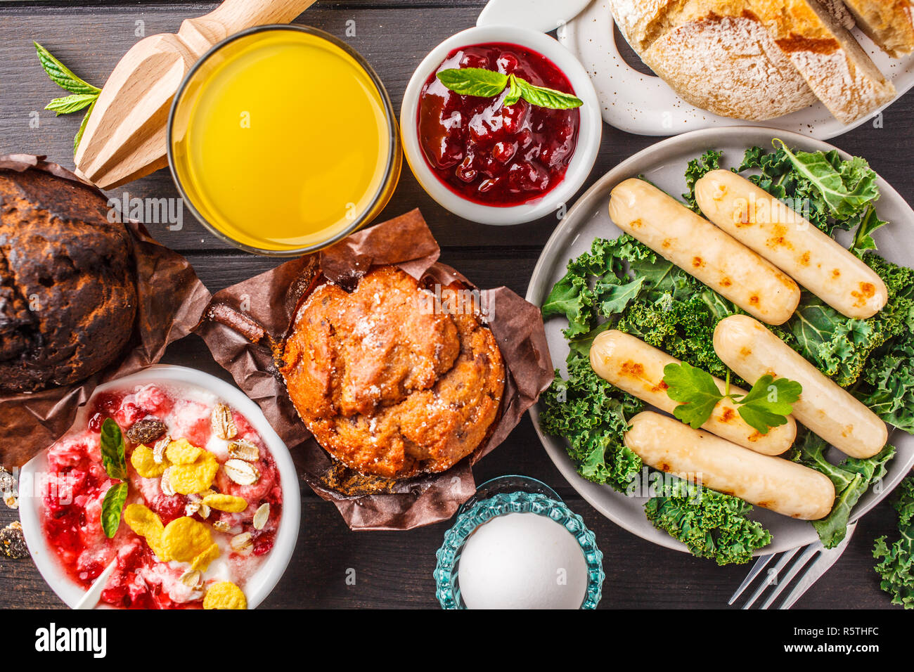 Desayuno con muffins, salchichas asadas, zumo, pan fresco y parfait sobre una mesa de madera oscura, vista superior. Foto de stock