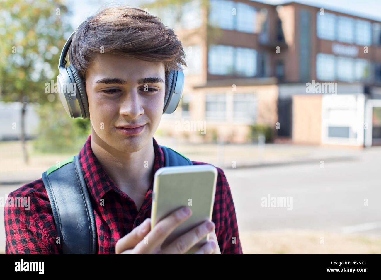 Adolescente masculino estudiante fuera del edificio del Colegio Streaming de música desde teléfonos móviles a auriculares inalámbricos Foto de stock