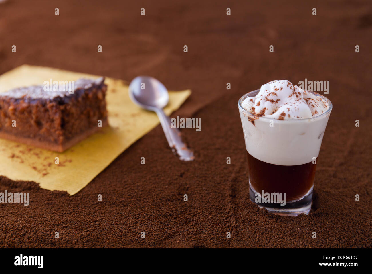 Poco taza de piccolo latte macchiato sobre una mesa cubierta con café molido como fondo y Brownie de chocolate con crema de cacao y virutas de coco Foto de stock