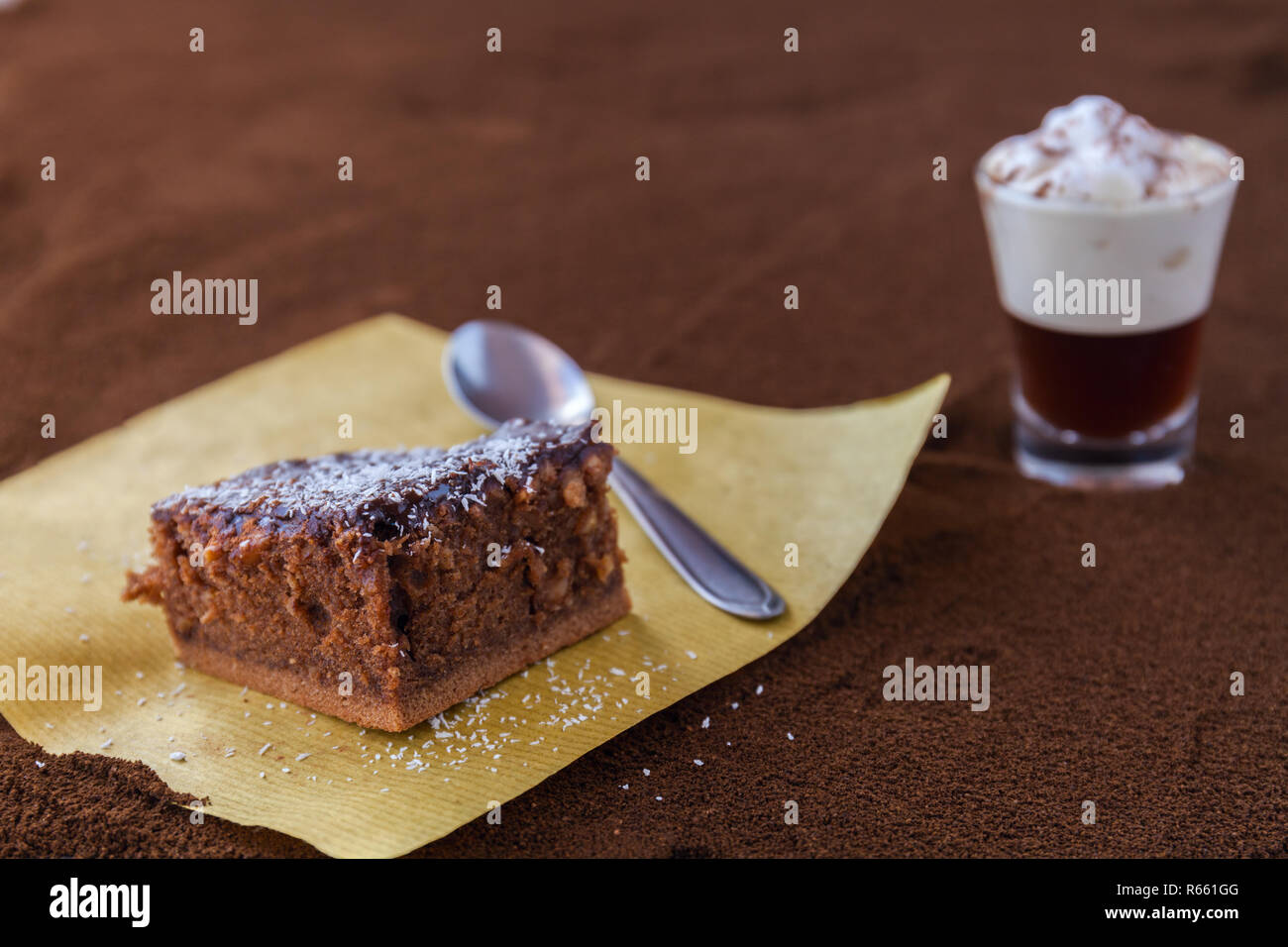 Poco taza de piccolo latte macchiato sobre una mesa cubierta con café molido como fondo y Brownie de chocolate con crema de cacao y virutas de coco Foto de stock