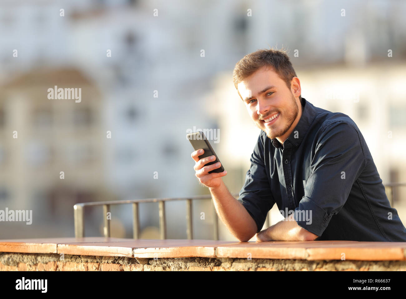 Feliz Hombre sujetando un teléfono inteligente mirando a la cámara en un balcón al atardecer Foto de stock