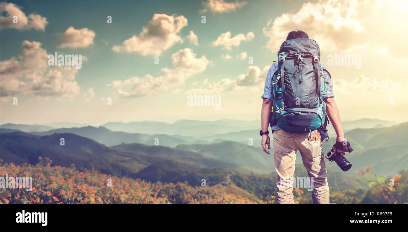Un muchacho joven con una mochila de viajero de pie en un acantilado, sosteniendo la cámara blanca y viendo las montañas nubladas. Estilo de vida fotos de viajes Foto de stock