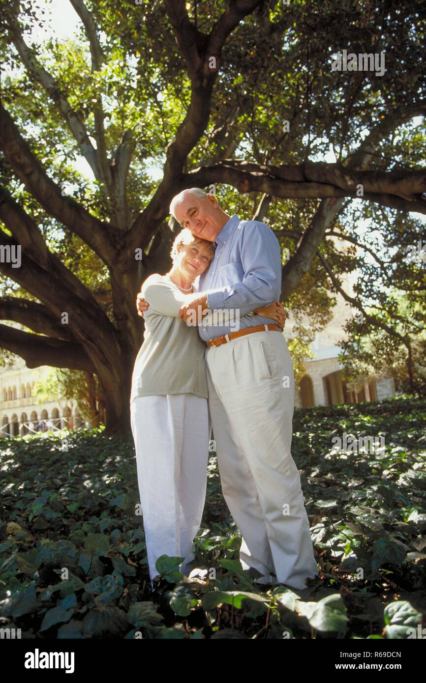 Ganzfigur, infierno gekleidetes Seniorenpaar steht Im Sommer zaertlich umarmt unter einem Baum Foto de stock