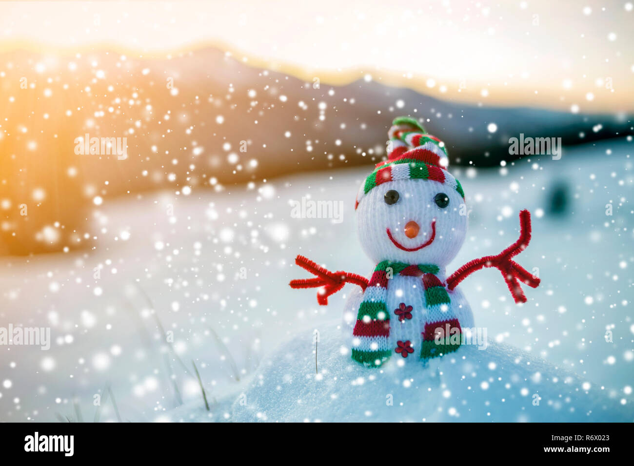 Pequeño bebé juguete divertido muñeco de nieve en el gorro de punto y  bufanda en nieve profunda piscina sobre el paisaje de las montañas borrosa  y caer copos grandes antecedentes. Feliz Fotografía