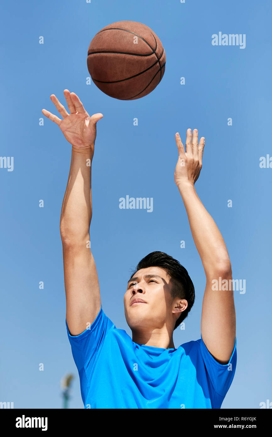 Joven jugador de baloncesto asiático haciendo un tiro libre con fondo de cielo azul. Foto de stock