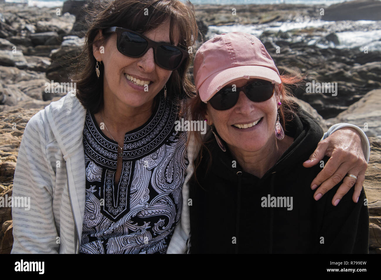 Dos mujeres caucásicas mujer sonriente baby boomers sentados juntos en las rocas a la orilla del océano Foto de stock