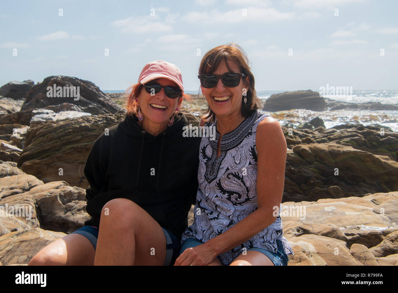Dos mujeres caucásicas mujer sonriente baby boomers sentados juntos en las rocas a la orilla del océano Foto de stock