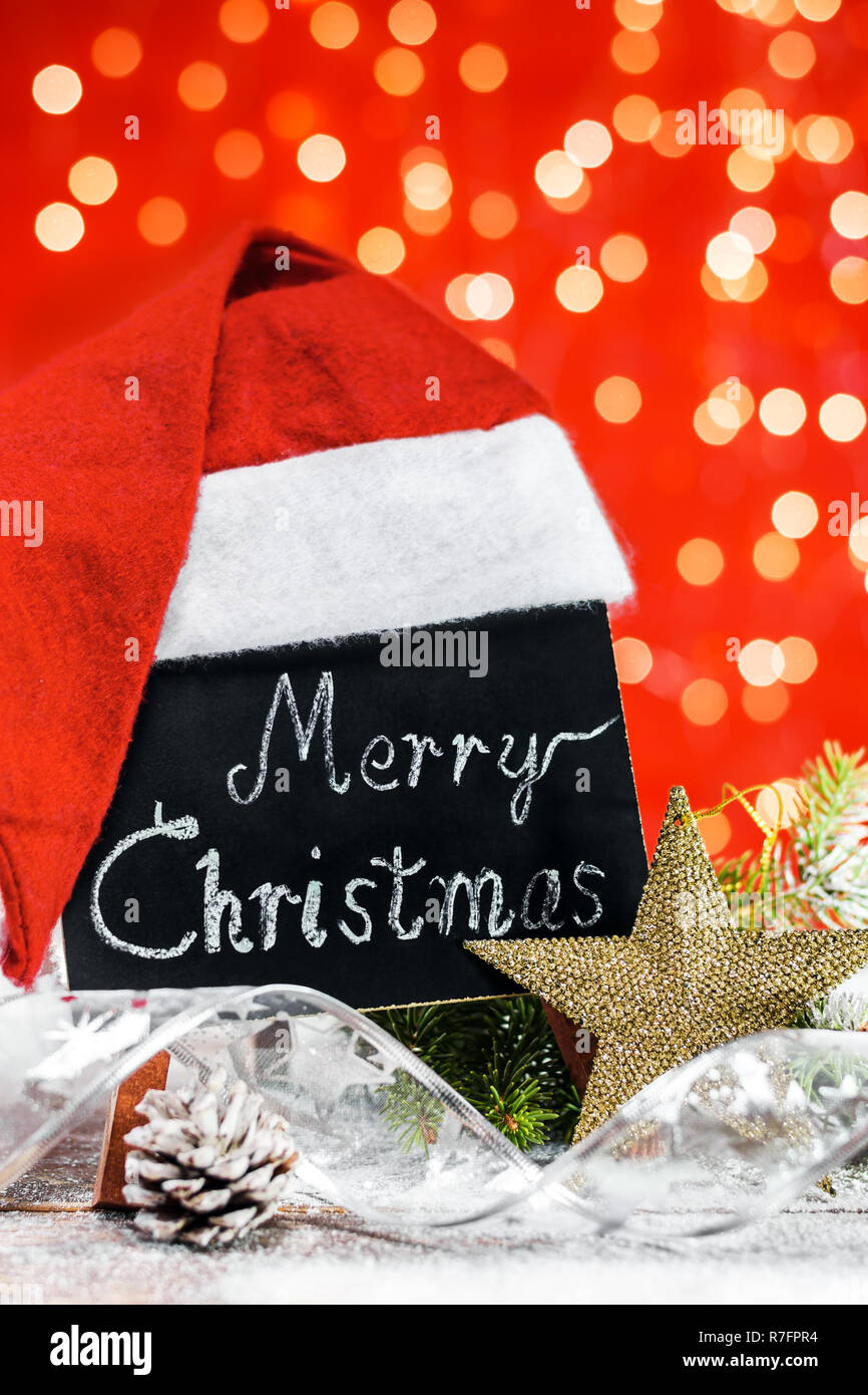 Un cuadro con el texto "Merry Christmas" escrito en una tiza cubierto con gorro de Santa Claus en una placa de madera cubierto con nieve con una rama de abeto, pinecone Foto de stock