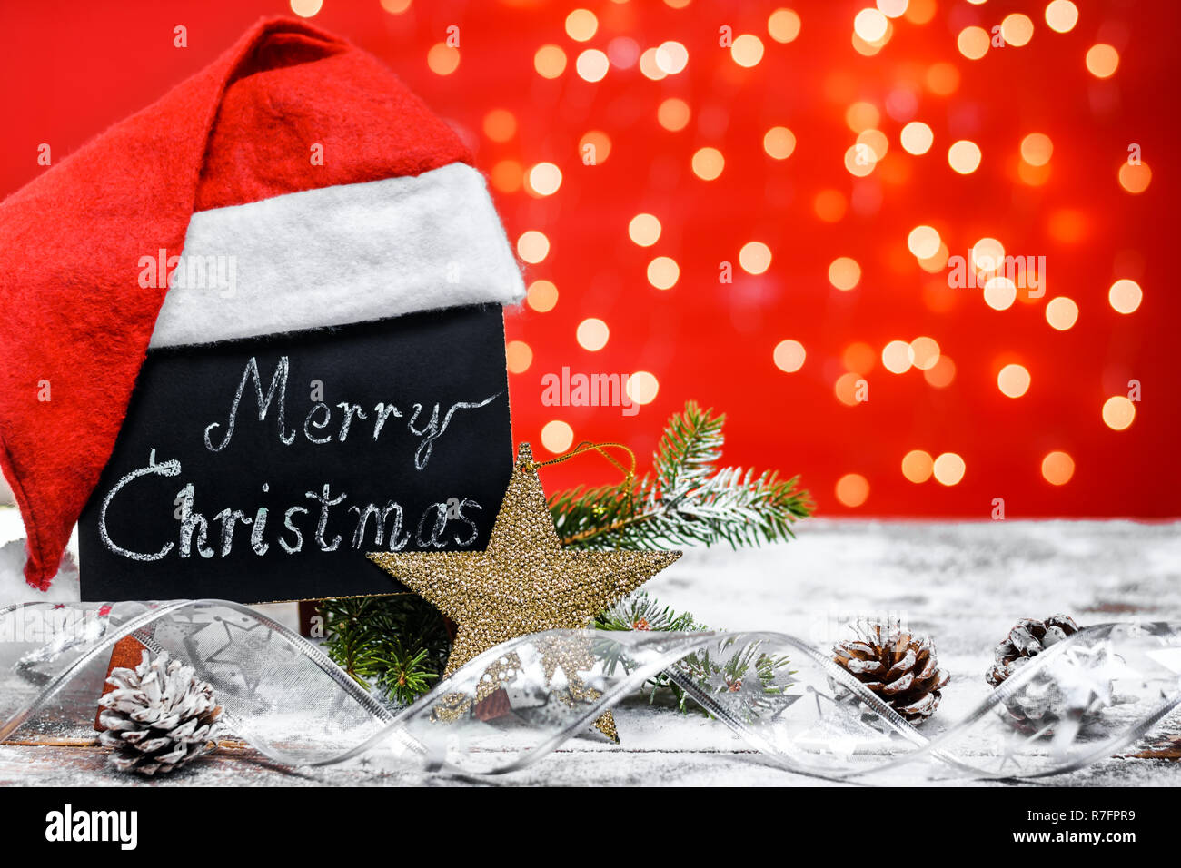 Un cuadro con el texto "Merry Christmas" escrito en una tiza cubierto con  gorro de Santa Claus en una placa de madera cubierto con nieve con una rama  de abeto, conos Fotografía