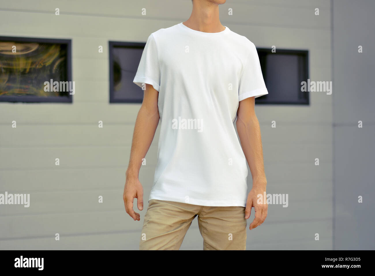 Plantilla urbana de ropa. Yong slim Guy en un blanco camiseta y pantalón  marrón permanezca cerca de la pared de perfilado de metal blanco con  ventanas negras. Boceto ca Fotografía de stock -