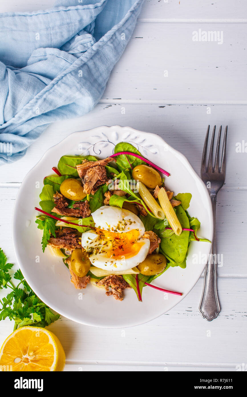 Ensalada de atún con pasta, aceitunas y huevo en una placa blanca sobre un fondo de madera blanca. Foto de stock