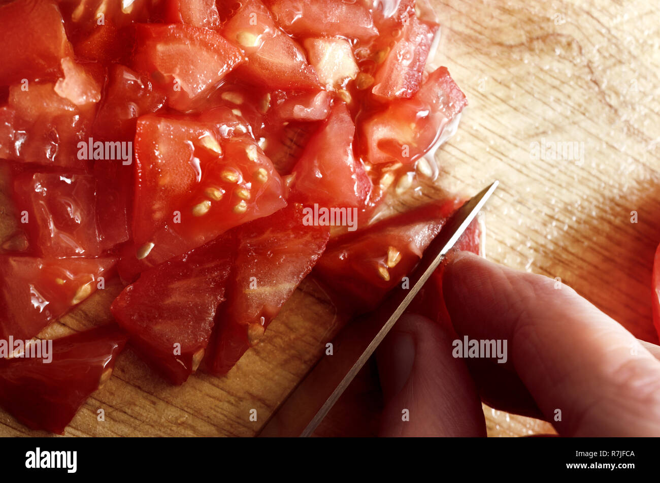Fotografía cenital de la mano que sujeta la cuchilla para picar y cortar jugoso, rojo tomate ensalada cruda sobre una tabla de cortar de madera. Foto de stock