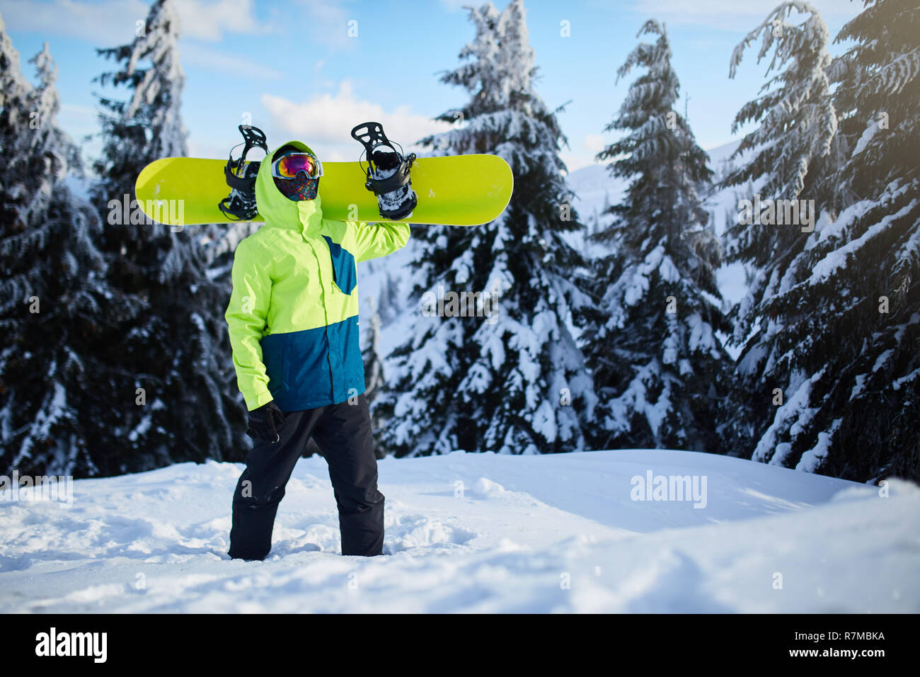 Snowboarder escalada de alta montaña llevando su snowboard a través del  bosque para travesías freeride y portando gafas reflectantes, colorida ropa  de moda y pasamontañas en estación de esquí. Deportes de invierno