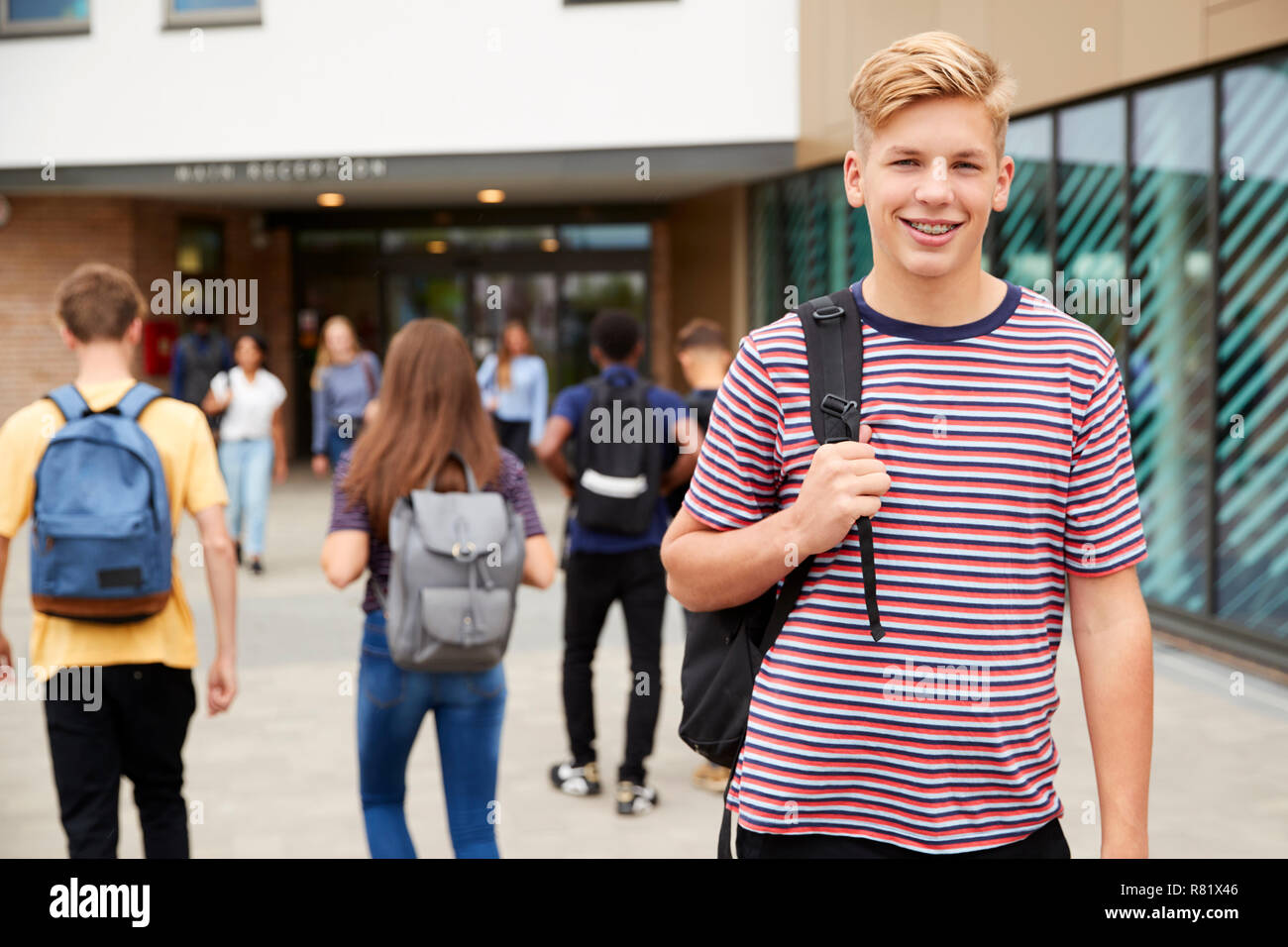 Retrato del estudiante de la High School secundaria masculina sonriente fuera del edificio del colegio con otros estudiantes adolescentes en segundo plano. Foto de stock