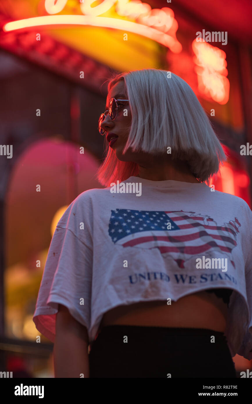 Retrato cinematográfico de la chica rubia con camiseta blanca y la bandera norteamericana en señal de neón en la noche Foto de stock