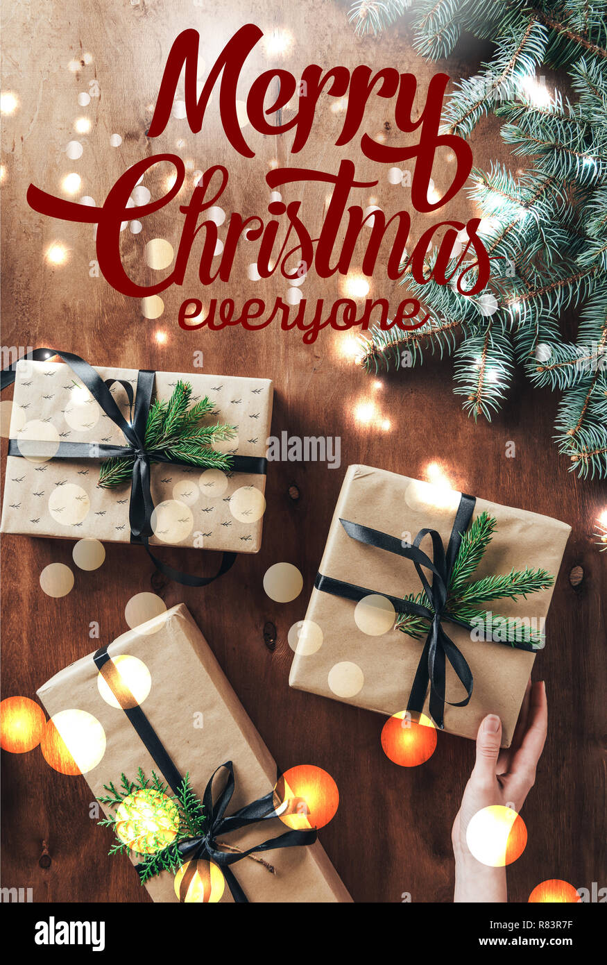 Vista recortada de mujer tocando cajas regalo sobre fondo de madera con ramas de árbol de navidad y "Merry Christmas everyone' rotulación Foto de stock