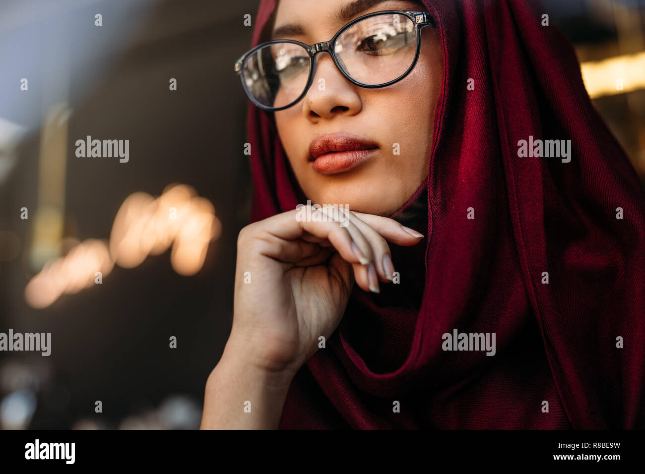 Close Up retrato de mujer joven llevar el hijab y anteojos con su mano en la barbilla apartar la mirada y el pensamiento. Mujer musulmana reflexiva mirando a otro lado. Foto de stock