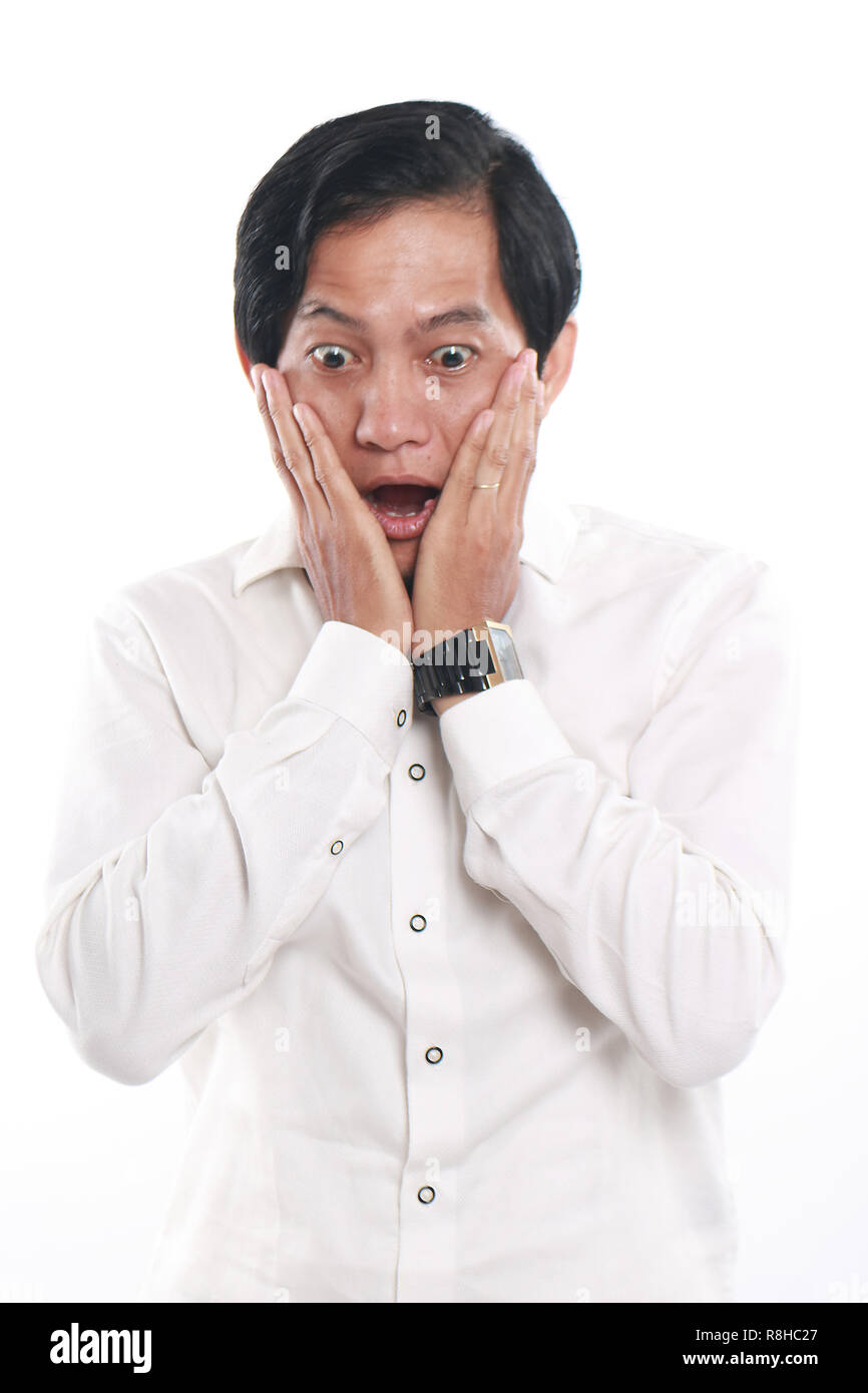 Imagen de Foto retrato de un joven gracioso empresario asiático conmocionado, cerca en vertical con gesto sorprendido, ambas manos en las mejillas, sobre fondo blanco. Foto de stock